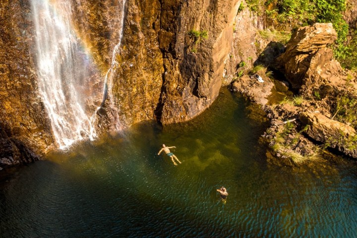 Two people swim near a waterfall with a visible rainbow.