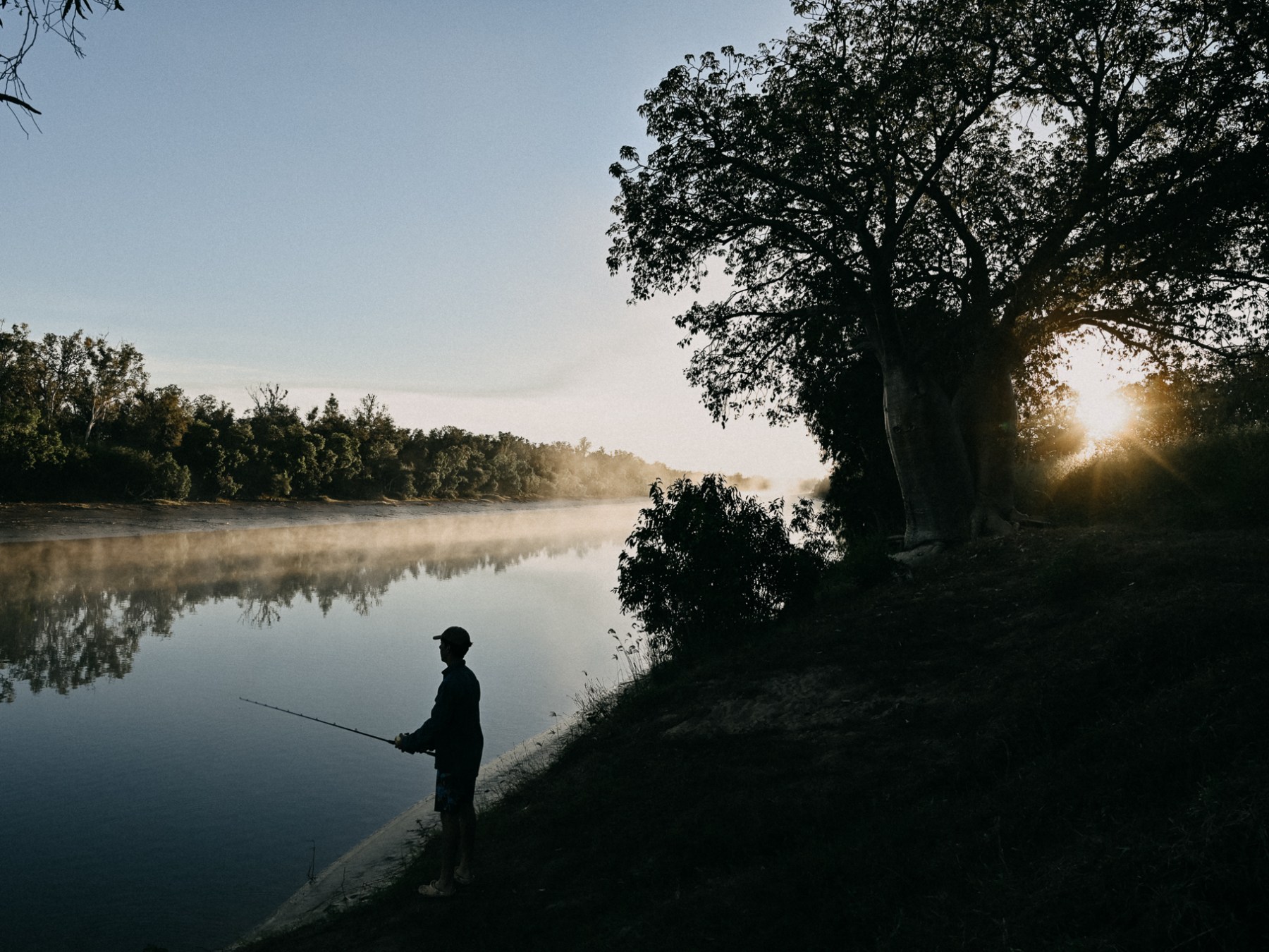 Person fishing by a calm river with trees, mist, and sunrise.