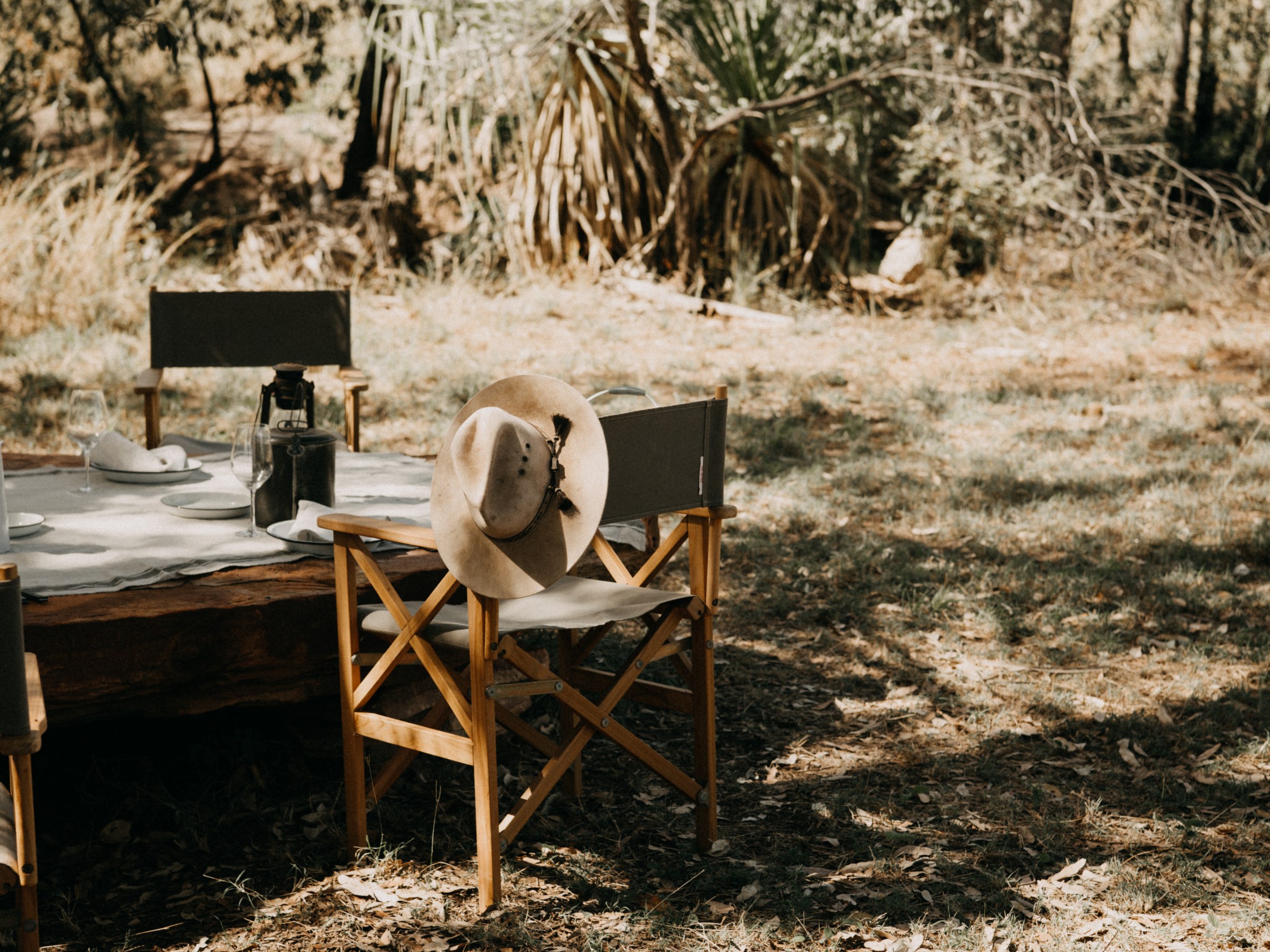 Outdoor table setting with wooden chairs and a hat on one chair, surrounded by dry grass and trees.