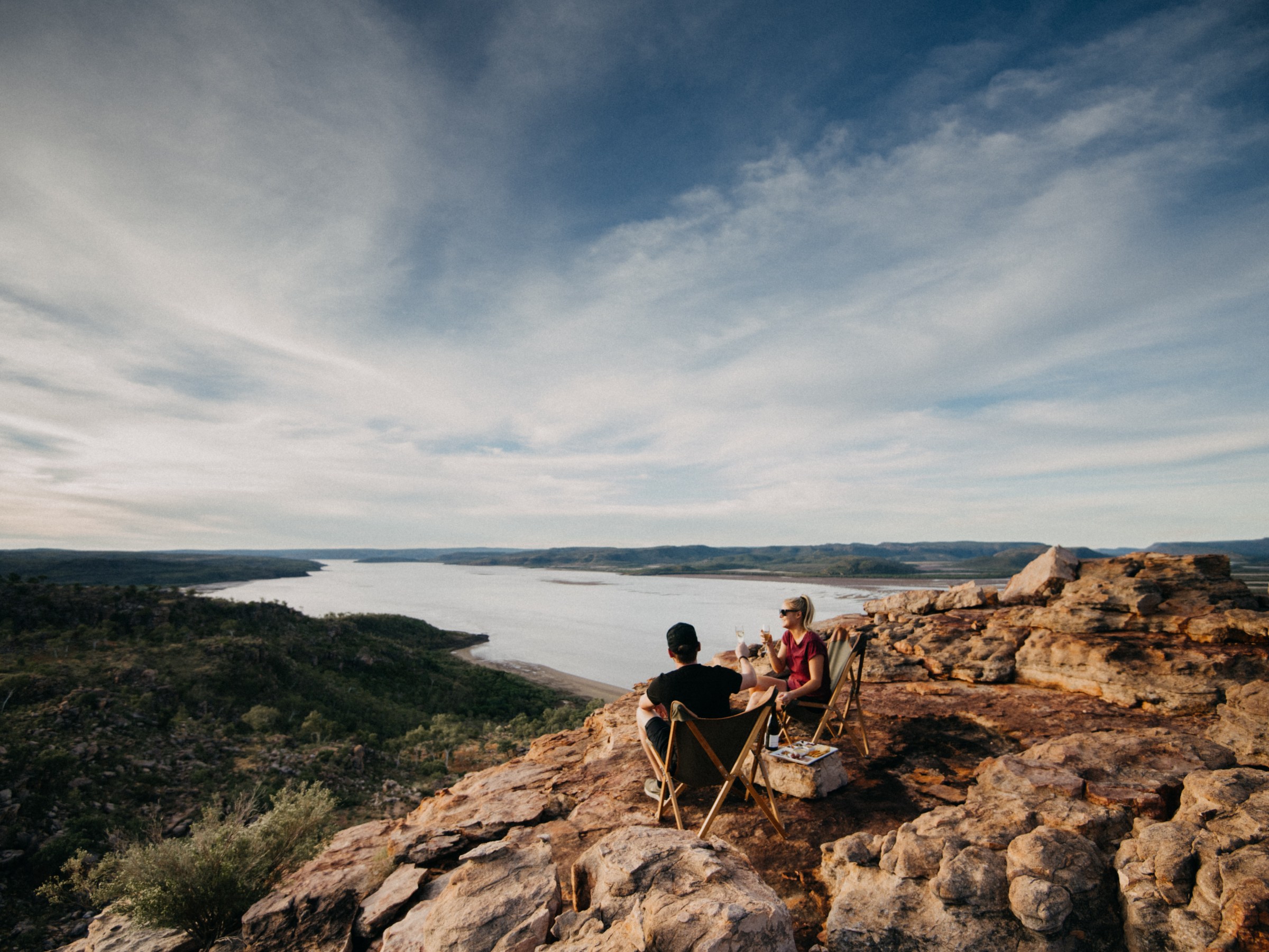 Two people sit on rocks overlooking a river and landscape.
