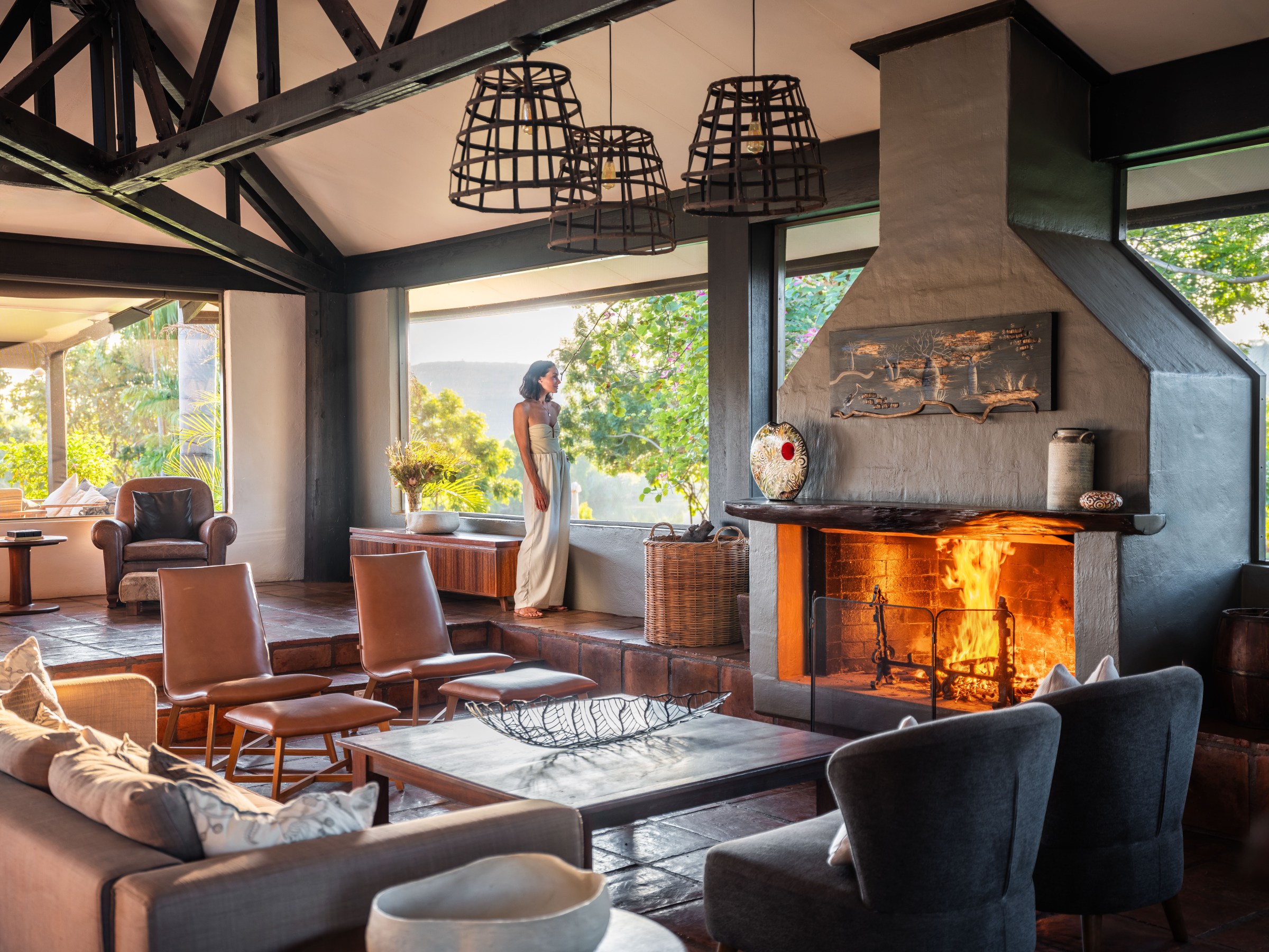 Cozy living room with a lit fireplace and a person standing by large windows.