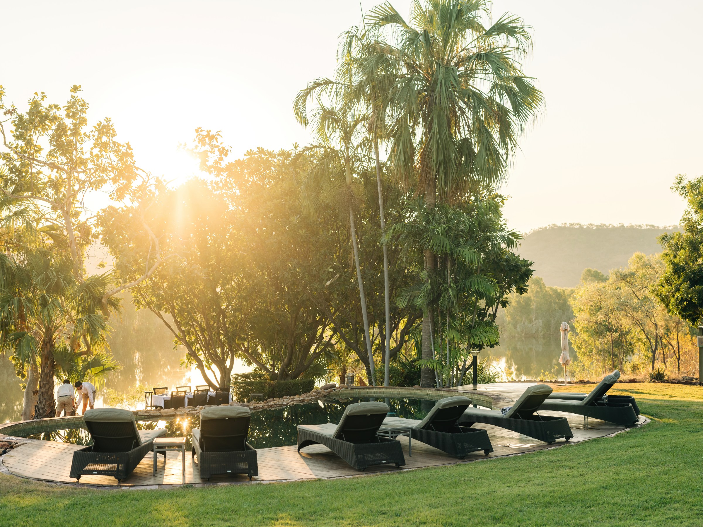 Sunlit pool with lounge chairs surrounded by trees and greenery.