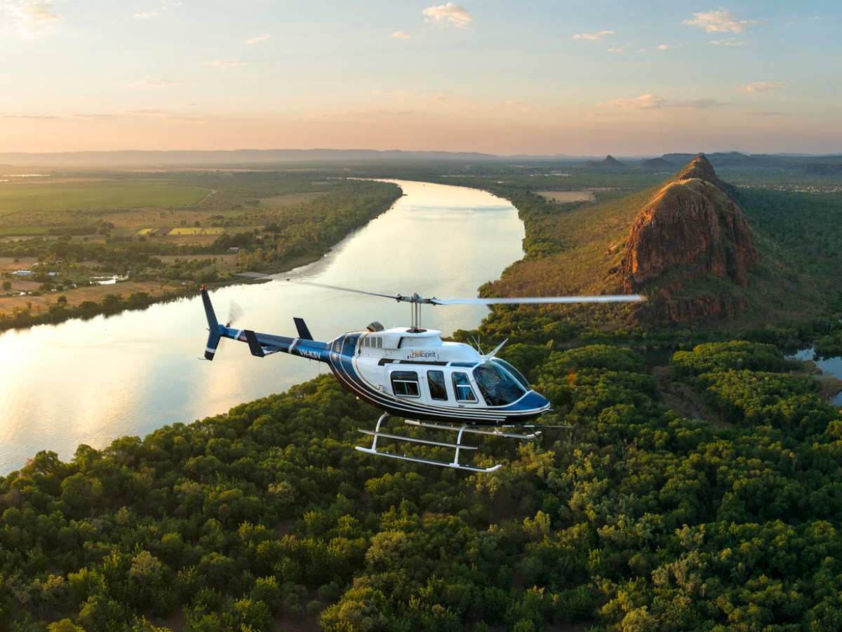 Helicopter flying over a river and forest at sunset with a hill in the background.