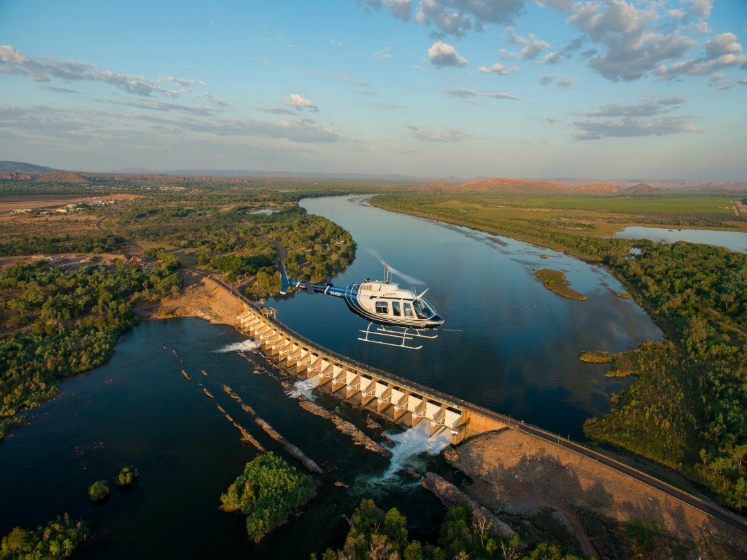 Helicopter hovering above a dam with river and landscape in view.