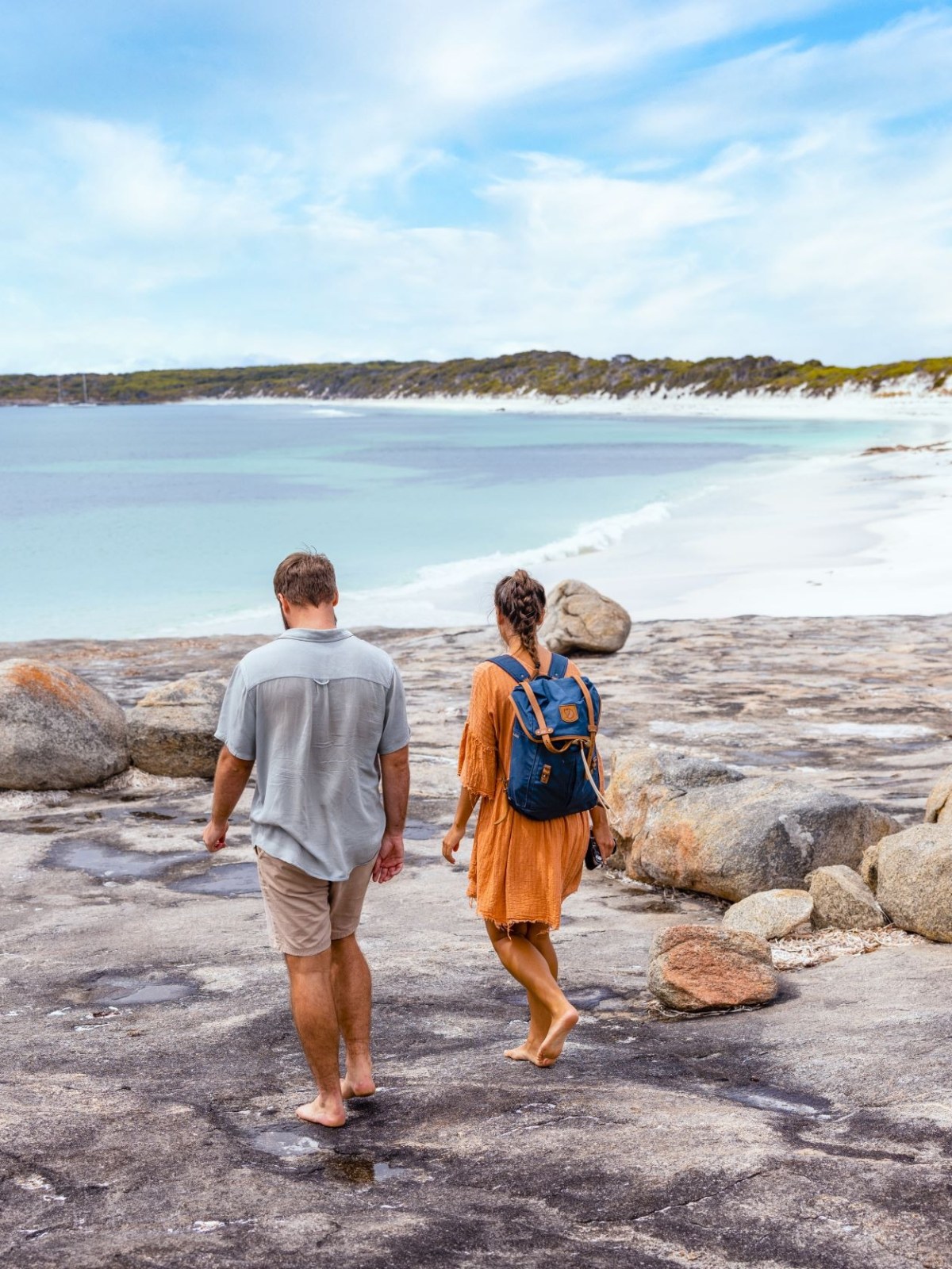 Couple walking on rocky shore towards a beach with clear skies.