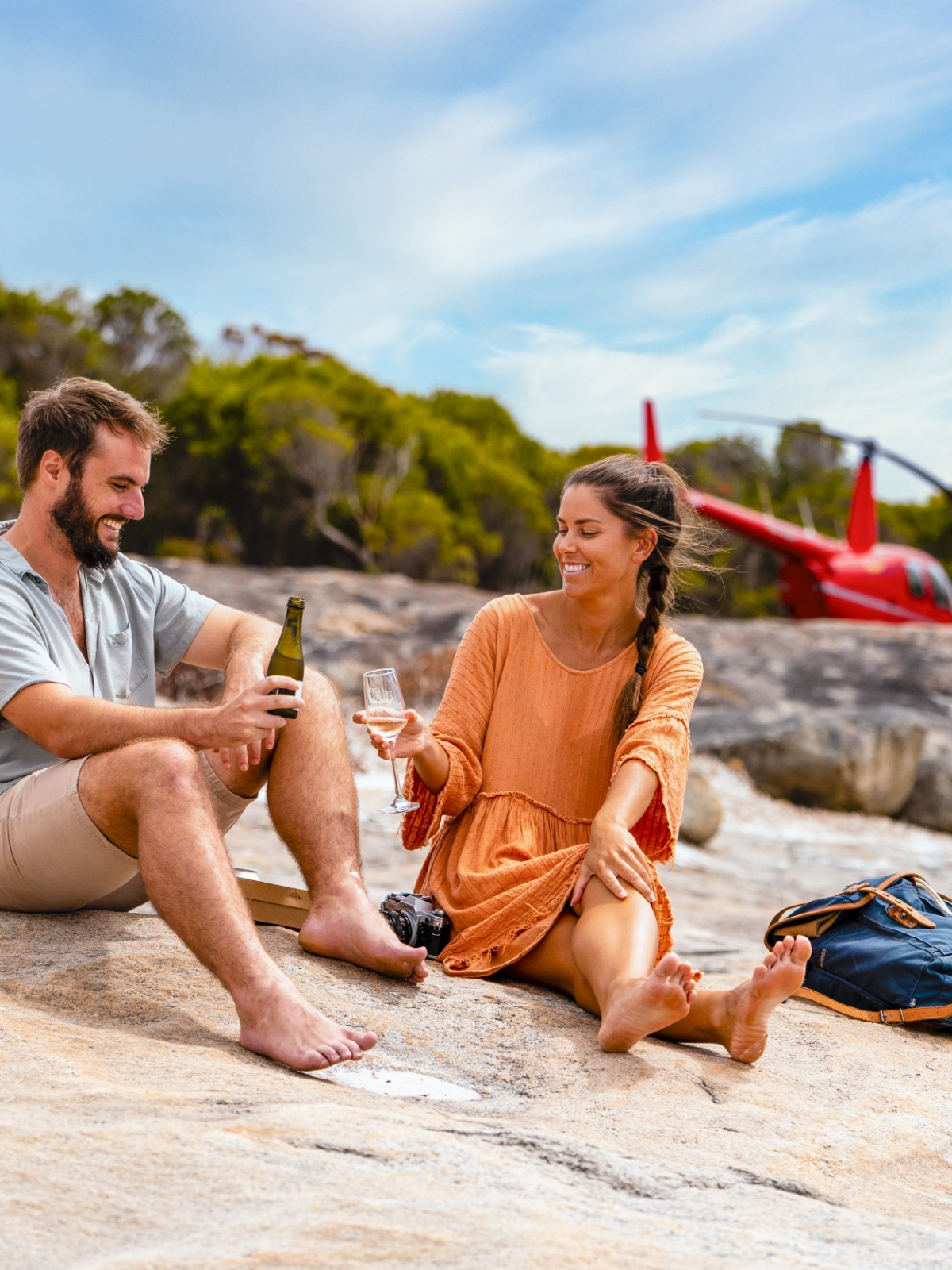 Couple sitting on rocks by a red helicopter and trees, holding drinks.