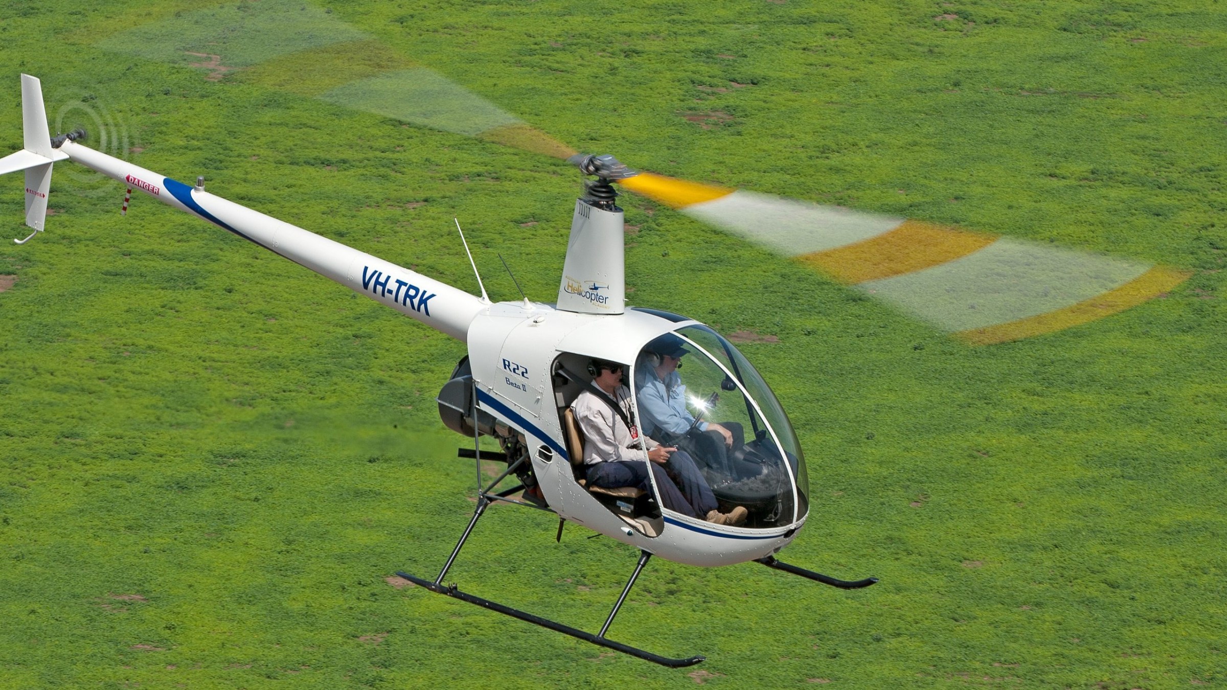 Small white helicopter flying over grassy field with two passengers.