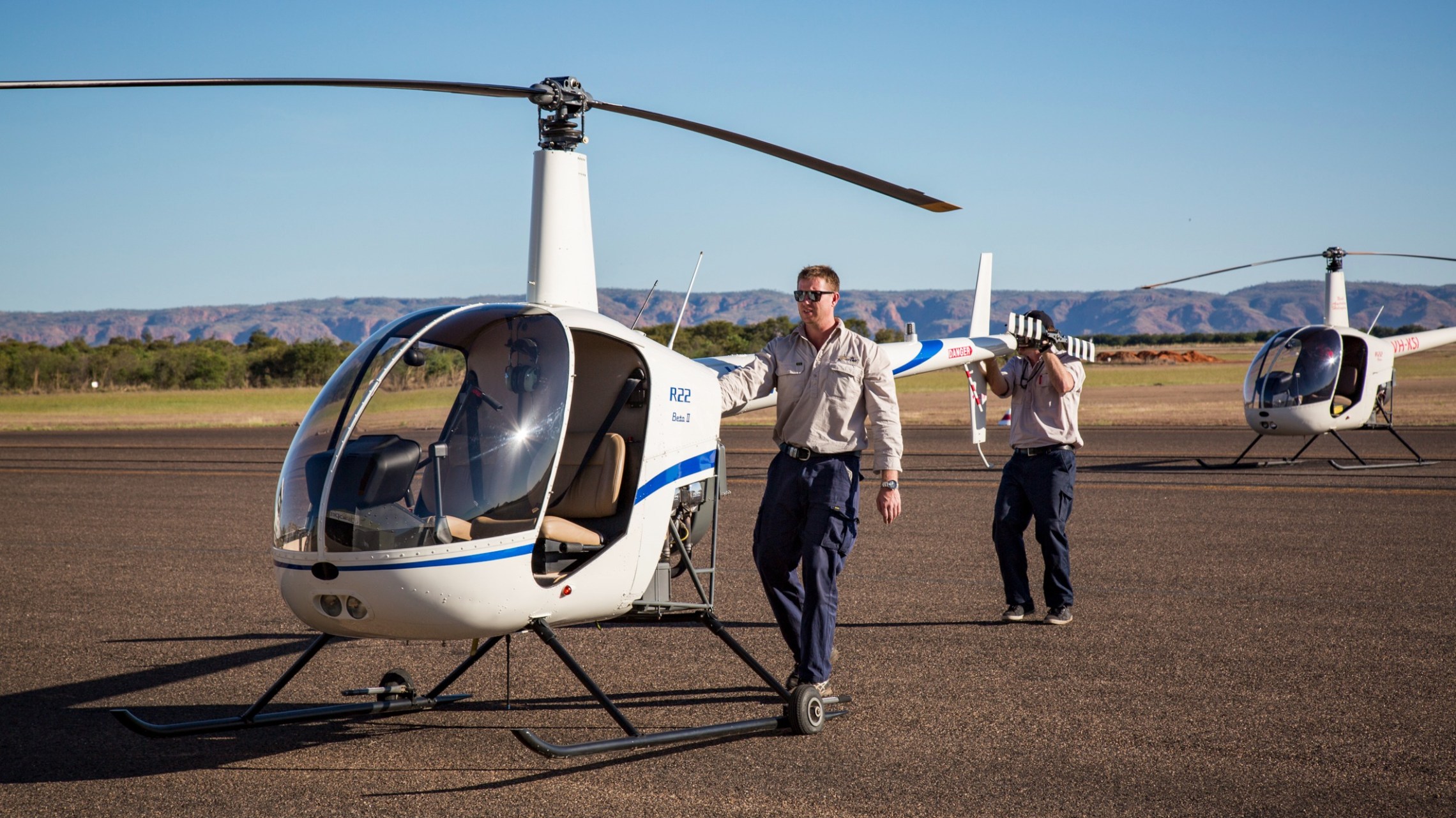 Two helicopters on runway with two pilots in uniforms beside them.